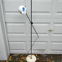 Full view of the vintage adjustable floor lamp placed outdoors against a white garage door. Shows white spherical lamp head, black metal adjustable arms, cream round base, and power cord.