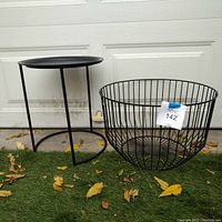 Front view of the black wire basket and small black metal end table side by side on artificial grass in front of a white garage door. Basket is large and open wire design, table is small with removable tray top.