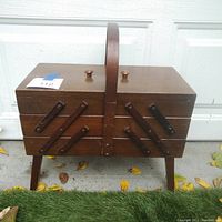 Closed view of the wood accordion sewing box showing wooden legs, handle, and two knobs on top with visible wear and scratches.