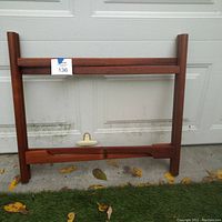 Photo of the folded teak table base standing upright against a garage door on green grass with dry leaves scattered around.
