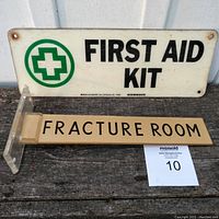 Photo showing both the First Aid Kit metal sign and the Lucite Fracture Room sign with attached mount on a wooden surface.