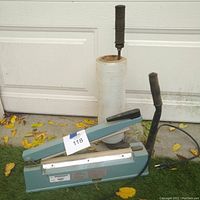Omcan heat sealer with black handles, teal body, and a partial roll of plastic on a vertical holder standing on grass outside a white garage door.