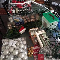 Photo showing multiple strings of Christmas lights, plastic ball ornaments in cardboard trays and bag, artificial greenery and gold berry sprays, Christmas stockings, holiday cards, and Snapware ribbon storage box
