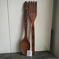 Full view of the tiki style wooden fork and spoon standing upright against a white background on a dark surface, showing overall shape and design.