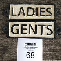 Pair of cast iron washroom plaques reading 'LADIES' and 'GENTS' photographed on wooden surface with auction label visible.