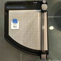 Full view of the X-Acto paper cutter showing the black base with a printed measurement grid surface and a vertical cutting arm equipped with blade holders.