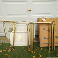Photo showing three removable TV tray tops with gold floral design and folding brass legs stacked separately, next to a cardboard box.