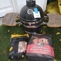 Overall view of the small ceramic egg-style smoker outdoors on grass. Shows black smoker with two wooden side shelves attached. Bag of small wood chips and bag of charcoal briquets placed in front for size comparison.