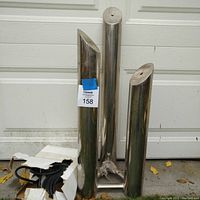 Front view of the stainless steel three-tier water fountain with pump and electrical wiring beside it. The fountain consists of three vertical cylindrical steel tubes arranged on a triangular steel base.