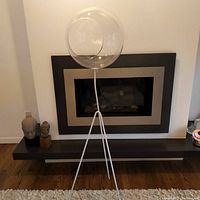 Full view of the terrarium standing nearly 5 feet tall in front of a fireplace. White metal tripod base supporting a large clear Lucite spherical globe.