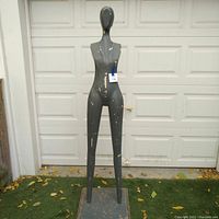 Full view of the full height female mannequin standing on base outside in front of white garage door. Visible paint chipping and weathering on mannequin body.
