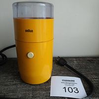 Full view of yellow Braun electric coffee grinder with power cord and white push button, sitting on wood surface