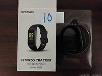 Fitness tracker inside packaging box next to the box on brown surface, showing front of box with product image and brand name Befitrack.