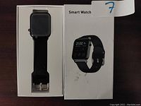 Black smart watch with silicone band in original packaging, displayed next to box showing image and product name.