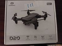 Front view of snow-white box labeled DeerC D20 Flying Drone showing an image of the black drone with four propellers and icons indicating altitude hold, 360 flip, trajectory flight, and 2.4 GHz.