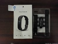 Front of the box showing the fitness tracker with black wristband and additional accessories in plastic packaging.