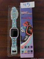 Kids smart watch in original packaging with colorful blue wristband featuring cartoon designs, displayed with product box showing key features.