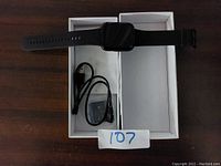 Smart watch lying across the inside of an open white box with a black USB charging cable and black wired earbuds in the box below it.