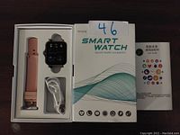 Smart watch, additional pink band, charging cable, and user guide in box. White box with smart watch branding and icons.