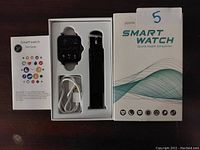 Photo of Souyie smart watch with display on, included user guide, charging cable, extra strap, all in original box showing packaging and contents.