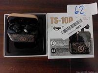 TS-100 earbuds inside black decorative charging case, shown with the product box included featuring the model name and Bluetooth 5.0 logo.