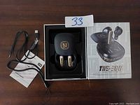 Photo of TWS-300 earbuds inside the charging case with USB charging cable and user manual placed beside the box showing product brand and model.