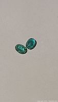 Two oval cut green emerald gemstones displayed together on a neutral background showing size and color match.