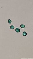 Five natural emeralds displayed on flat light background, showing the green color and brilliance of each stone.