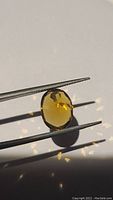 Close-up photo of natural oval Madeira citrine held by tweezers showing deep cognac color and faceted surface under direct light.