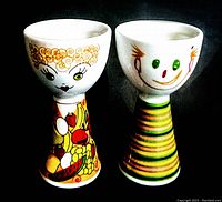 Front view of two mid-century ceramic egg cups, one with fruit and vegetable lady design and the other with hungry breakfast man design.