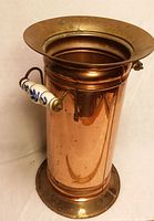 Front side view of the tall copper cylindrical umbrella stand with one white and blue Delft ceramic-handled arm visible.