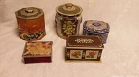 Five assorted antique English tin containers arranged on a light fabric surface in a single grouping, displaying varying shapes, colors, and decorative designs.