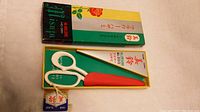 Photo of the white-handled scissors with red blade cover inside the open original box showing Japanese text and floral decoration.
