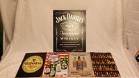 Five metal signs displayed together on a white backdrop. Signs include Jack Daniels, Guinness, Canada Dry, and other whiskey bottle designs. Signs are rectangular and arranged with one circular sign visible. All appear in good condition with no visible damage.