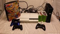 White PS3, black PS3 with skin, Xbox 360 console, three controllers, cables, Xbox game discs