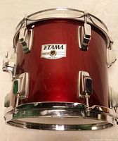 Close-up photo of Tama tom drum showing red lacquered shell with chrome hardware and Tama badge