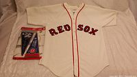 Photo showing the Boston Red Sox vintage jersey laid flat with the unopened set of three mini baseball pennants to the left side.