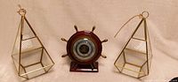 Two glass and brass hanging air plant holders in reverse diamond shapes flanking a nautical themed barometer designed as a ship's wheel, arranged on a beige cloth background.