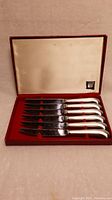 Six Oneida steak knives with serrated blades and textured silver-tone handles in original red box with beige satin lining inside lid.