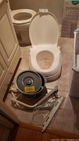 Full view of white plastic commode seat with open lid, grey pail and lid, and assembly hardware on tiled floor.