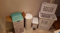 Set of four items: Doona PurePail diaper pail with box, white plastic garbage can with foot pedal, and two stacked white plastic laundry baskets.