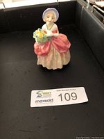 Full view of the Royal Doulton Cassie figurine showing the lady in a bonnet and pink sash holding a flower basket.
