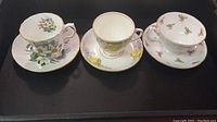 Three vintage fine bone china teacup and saucer sets displayed side by side showing floral hand-painted designs and gold rims.