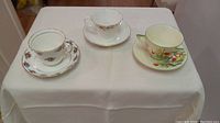 Three vintage fine china teacup and saucer sets displayed side by side on a white cloth.