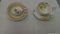 Two fine china teacup and saucer sets placed side by side on a table, showing floral designs and gold accents.