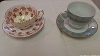 Two teacup and saucer sets on a white surface, left is floral pink gold trimmed, right is gray and white with gold trim