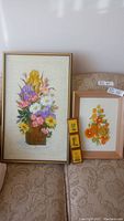 Two framed hand embroidery pictures displayed on a sofa, one large and one smaller, along with small yellow boxes.