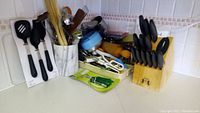 Full view of kitchen counter with knife block, utensils in holders including wooden spoons, spatulas, rolling pin, grater, and other kitchen tools scattered and organized.
