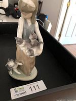 Front view of the Lladro girl figurine holding three cats, with an additional cat at her feet, displayed in a black tray.