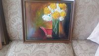 Frontal view of original Tom Brown Wilson '64 framed oil painting with white and yellow flowers in a green vase and red decorative piece, placed on a patterned sofa.
